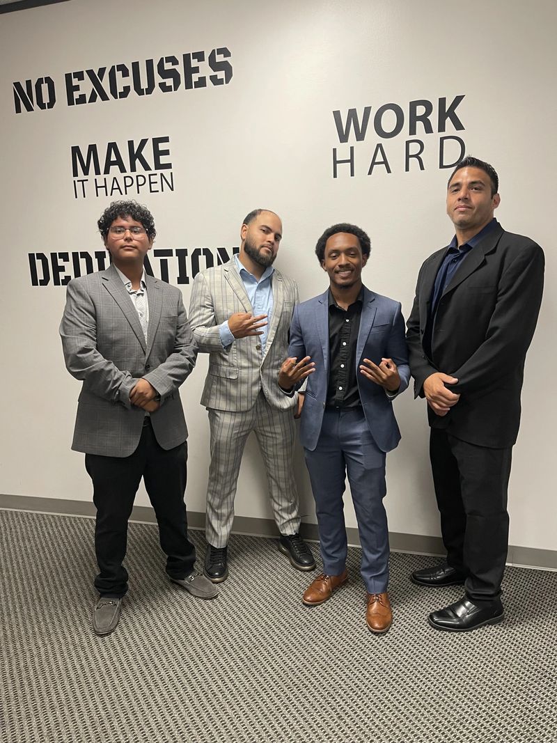 Four Mission III Management team members in front of a “Make It Happen — Work Hard” wall